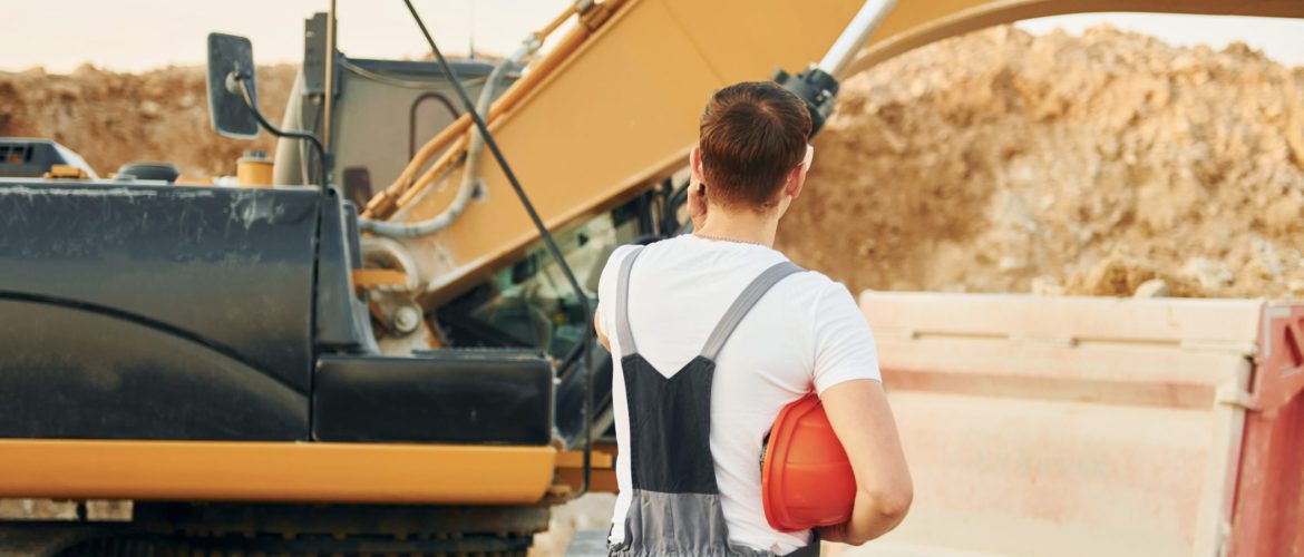 Standing against yellow vehicle. Worker in professional uniform is on the borrow pit at daytime.