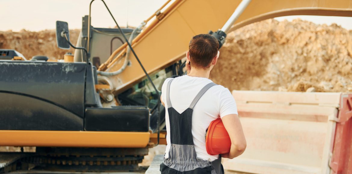 Standing against yellow vehicle. Worker in professional uniform is on the borrow pit at daytime.