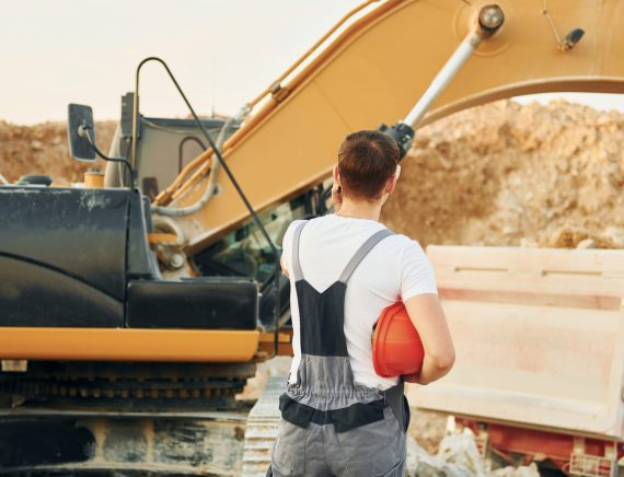 Standing against yellow vehicle. Worker in professional uniform is on the borrow pit at daytime.